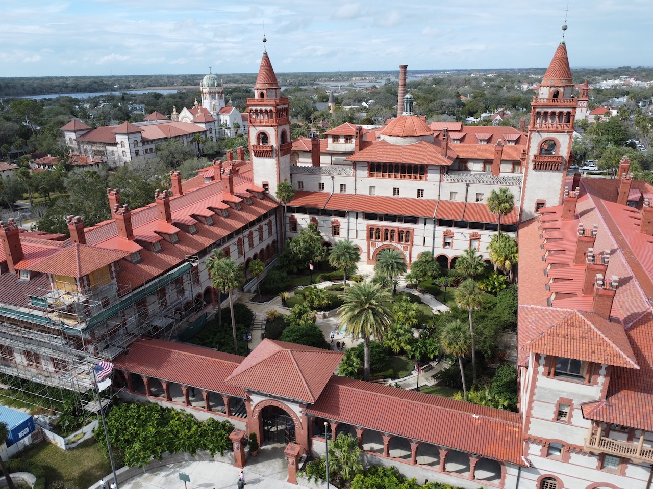 Vista aérea do Flagler College, edifício histórico em St. Augustine com telhados vermelhos e várias torres, rodeado por jardins bem cuidados e palmeiras, com outros edifícios visíveis ao fundo.